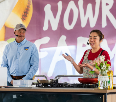 Mama Kim in a red apron speaks and gestures while standing behind a cooking station.